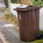 Brown wheelie bin on pavement beside a white picket fence