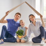 Man, woman and child sitting on the floor smiling and holding a cardboard roof over their heads