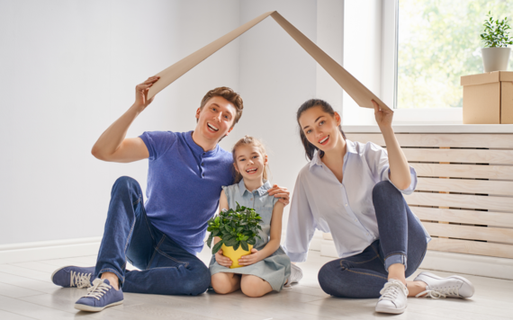 Man, woman and child sitting on the floor smiling and holding a cardboard roof over their heads