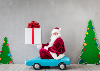 Person in father christmas outfit sitting on a child sized blue car holding a parcel with a red bow on top